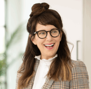 A woman with long brown hair and glasses, wearing a plaid blazer and a white ruffled blouse, smiles while looking slightly to the right in a bright indoor setting.